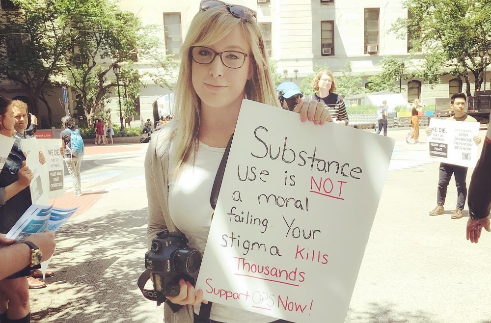 Brittany Salerno stands outside holding a sign that says, "Substance use is not a moral failing. Your stigma kills thousands. Support OPS now."